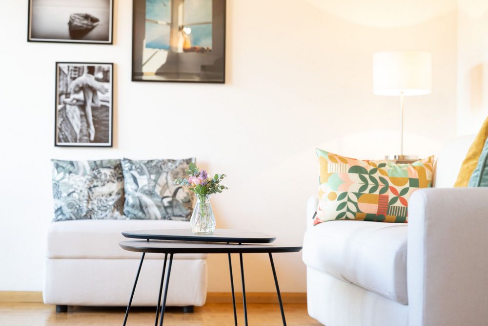 Heller Wohnbereich der Ferienwohnung Sonnenburg mit weißen Sofas, bunten Kissen, Couchtischen und einer Vase mit frischen Blumen.