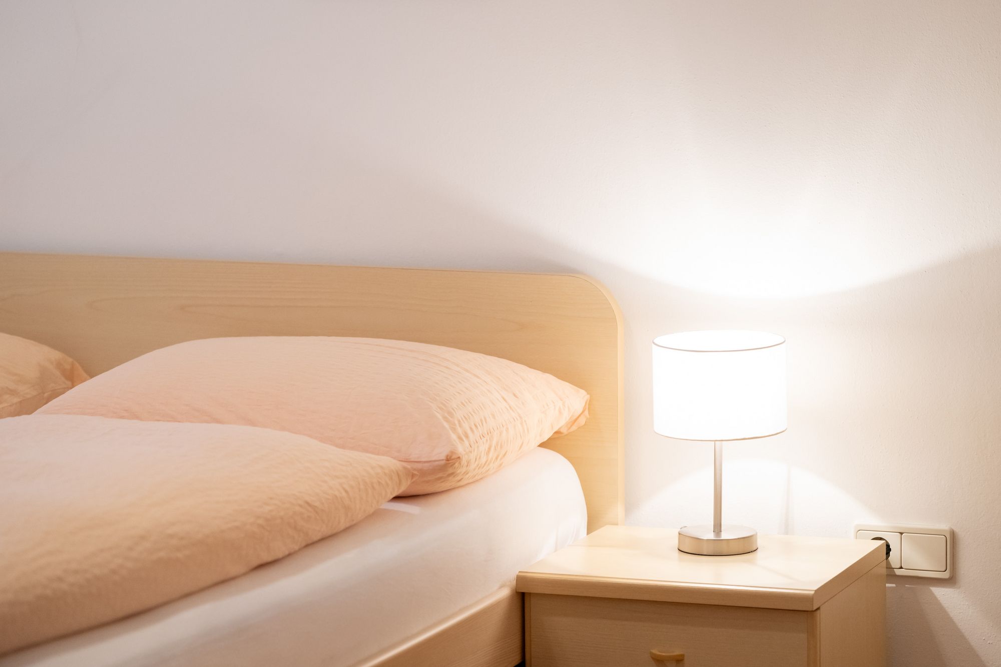 Detailaufnahme eines gemütlichen Bettes mit heller Bettwäsche, Holz-Nachttisch und eingeschalteter Leselampe in der Ferienwohnung Sonnenburg.