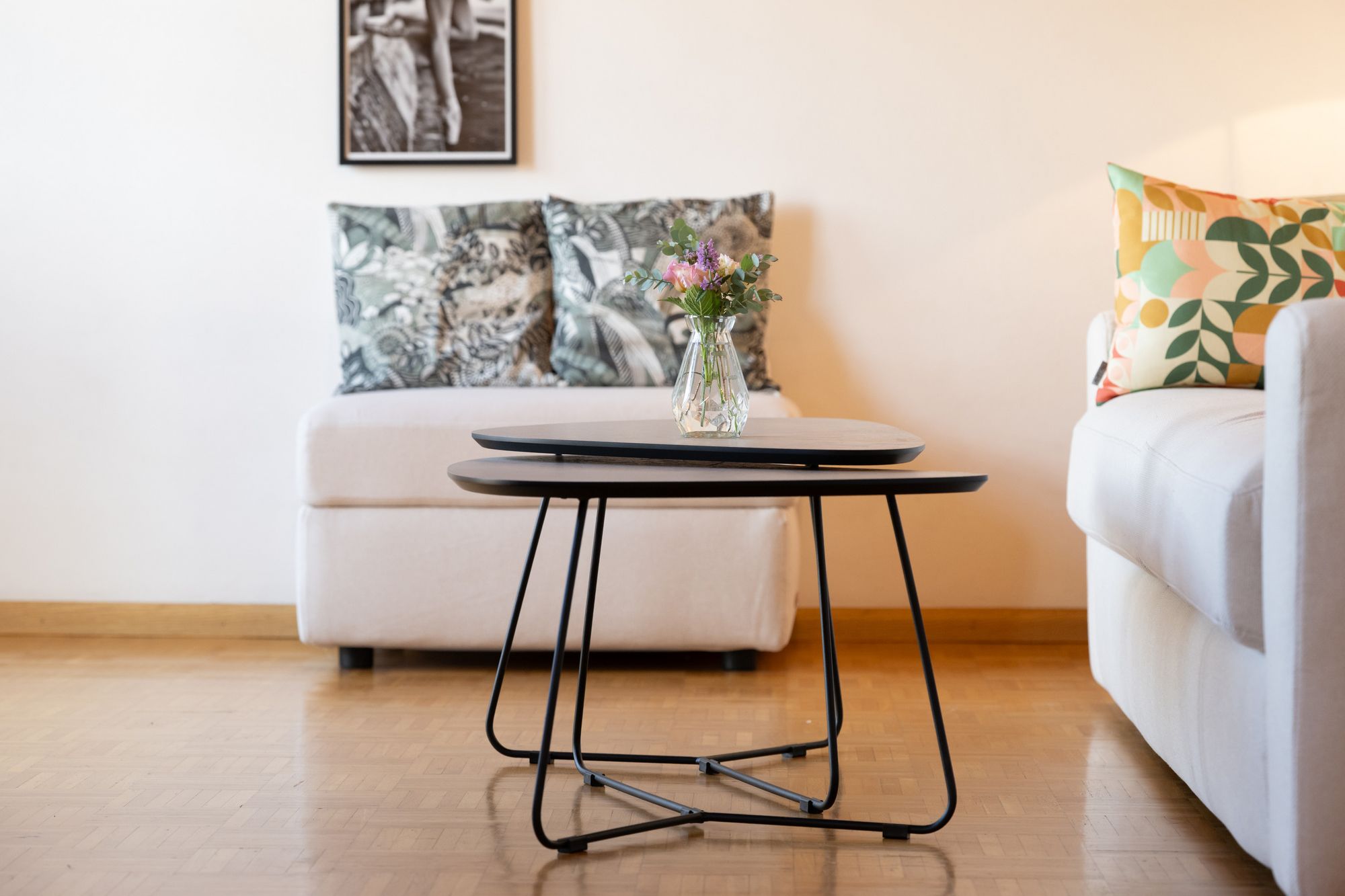 Heller Wohnbereich der Ferienwohnung Sonnenburg mit weißen Sesseln, bunten Kissen, zwei Couchtischen und einer Blumenvase.
